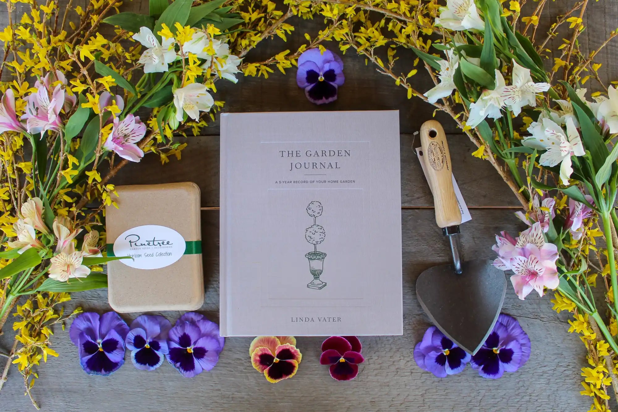 A garden journal surrounded by pansies, pink lilies, yellow forsythia branches, and a small garden trowel.