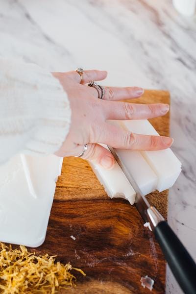 Hand and knife cutting soap base bar on a wooden cutting board.